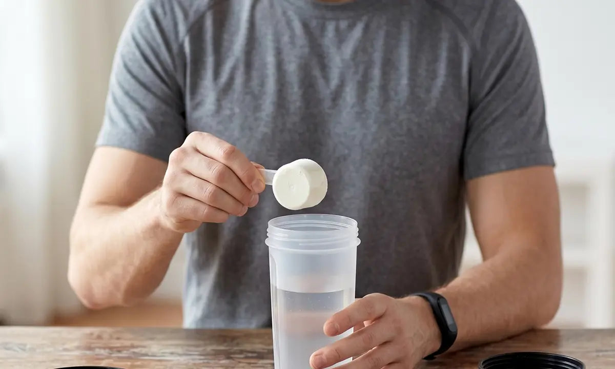 Homem de camiseta cinza segurando uma shakera com água e segurando um scoop de creatina encima do recipiente.