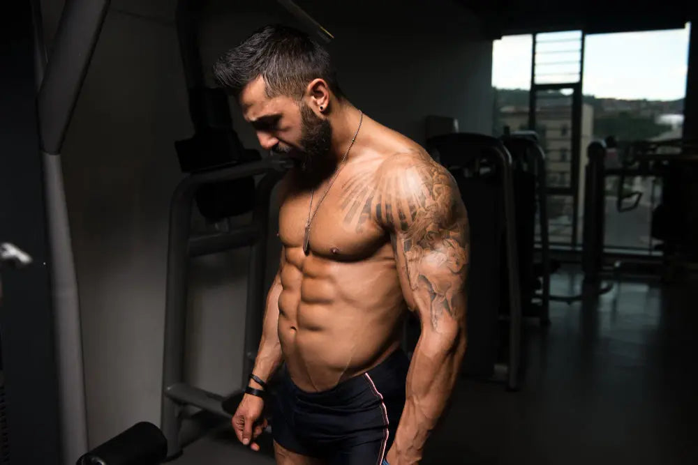 Homem com físico musculoso e definido em academia, observando o próprio corpo durante o treino de musculação.