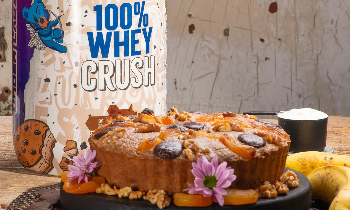 Bolo caseiro com frutas secas e nozes sobre uma tábua, acompanhado de bananas e um pote de whey protein ao fundo.