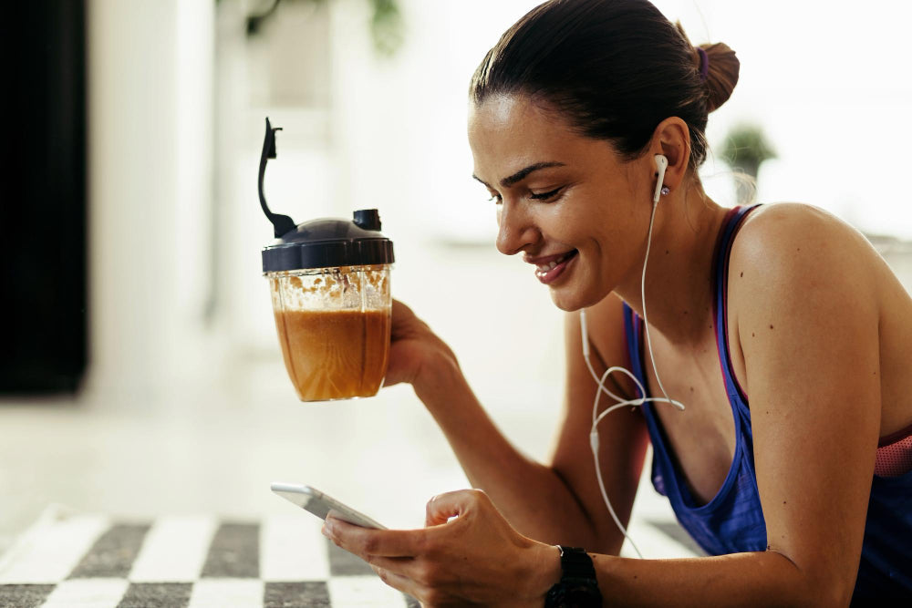 A imagem mostra uma mulher sorridente vestindo roupa de treino, ouvindo música com fones de ouvido e segurando um copo de shake ou smoothie.