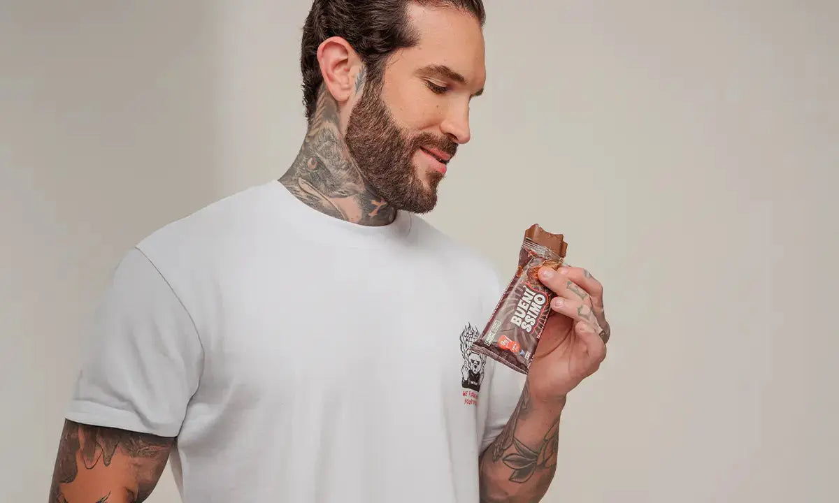 Homem com barba e tatuagens, vestindo camiseta branca, observa uma barra de proteína de chocolate que segura na mão, em um fundo neutro e iluminado.