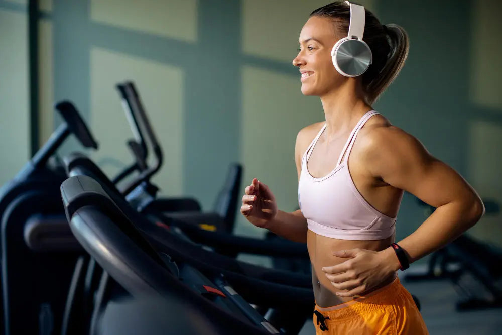 Mulher correndo na esteira em uma academia, usando fones de ouvido e roupa esportiva, representando a prática de treino de cardio.