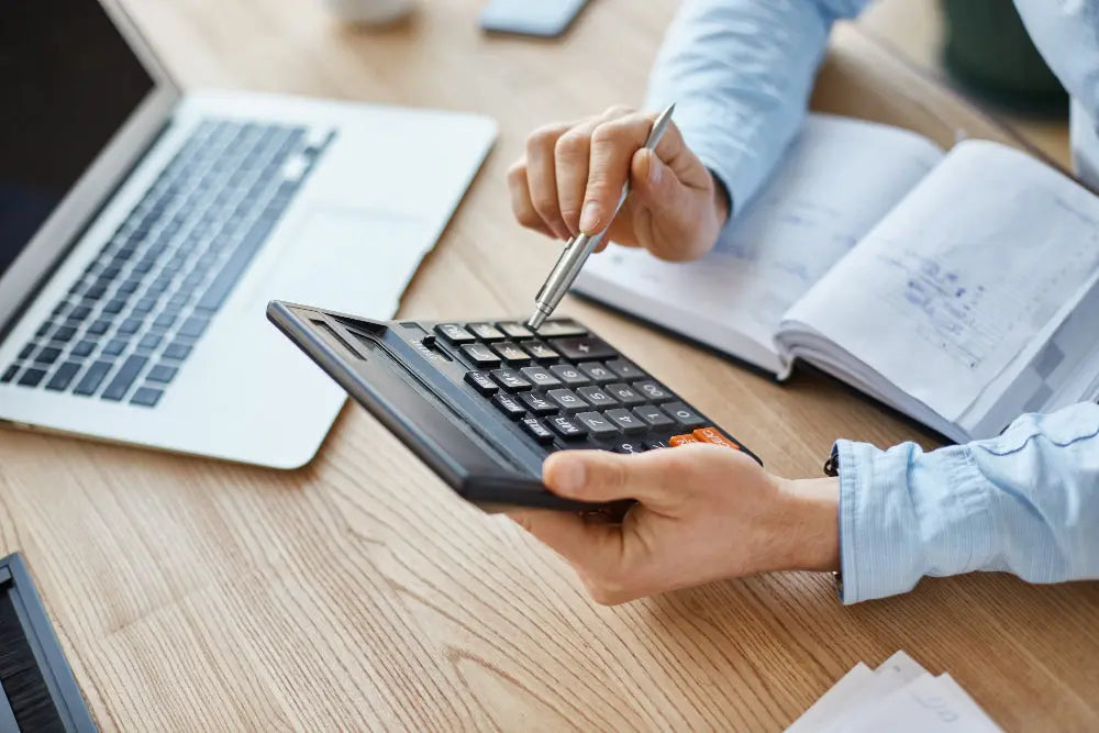 A imagem mostra uma pessoa usando uma calculadora enquanto segura uma caneta. Sobre a mesa há um notebook aberto e um caderno com anotações, indicando um ambiente de trabalho ou estudo focado em atividades financeiras ou contábeis.