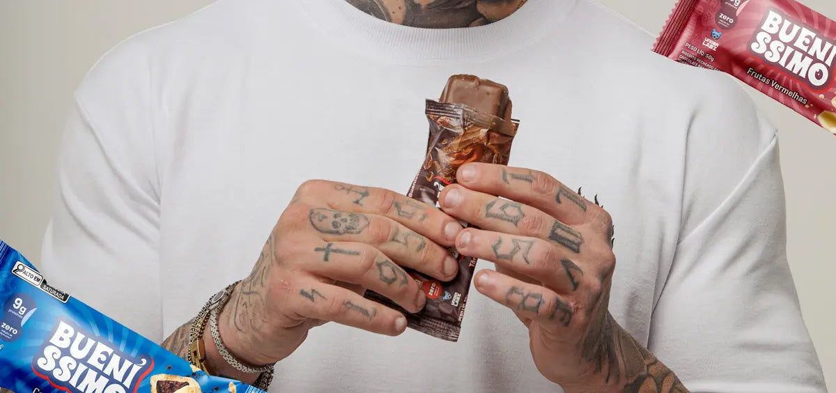 Homem de camiseta branca, com mãos tatuadas, abrindo uma barra de proteína Bueníssimo sabor chocolate, com embalagens do produto ao redor da imagem.