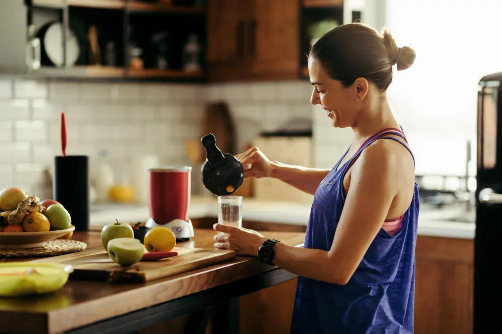 Uma mulher prepara um suco na cozinha, sorrindo, com frutas e um liquidificador sobre a bancada.