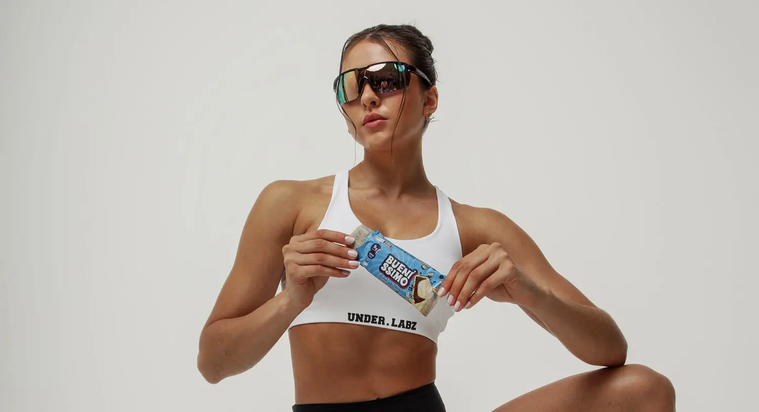 Mulher com look esportivo e óculos escuros segura uma barra de proteína ‘Bueníssimo’. Com visual confiante e estiloso, ela representa atitude e performance em um cenário clean e moderno.