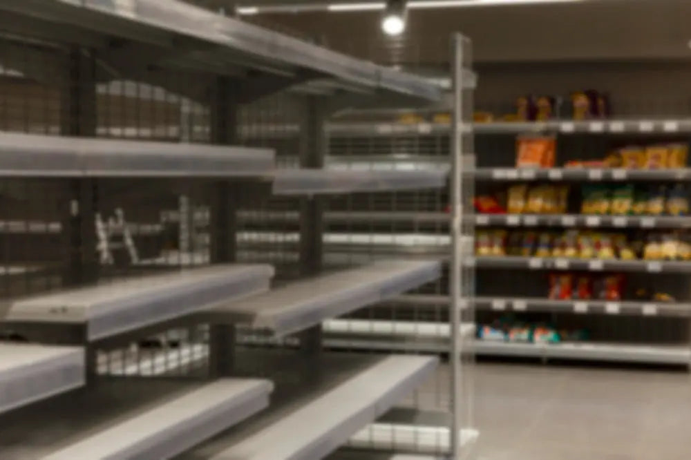 A imagem mostra prateleiras de mercado com espaços vazios e alguns produtos visíveis ao fundo. O ambiente é moderno, mas a foto está desfocada, sugerindo escassez de itens.